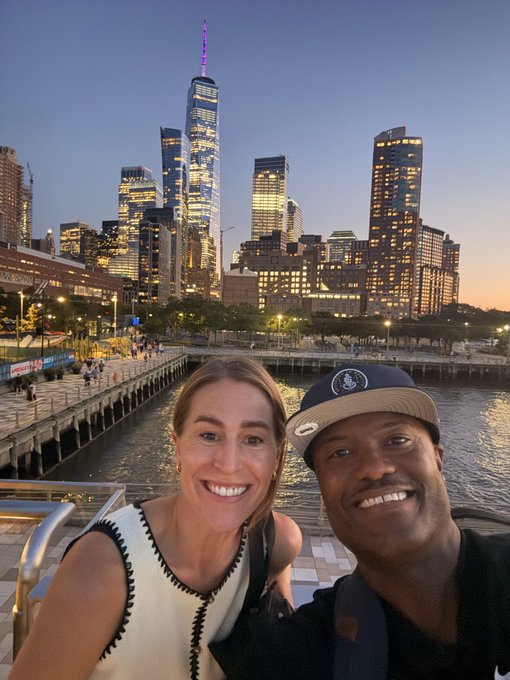 A smiling couple takes a selfie along the Manhattan waterfront at dusk, with the illuminated One World Trade Center towering prominently in the background, capturing their excitement for the Mainnet 9.16 launch as a relaxed, scenic cityside moment with @bobbilee.