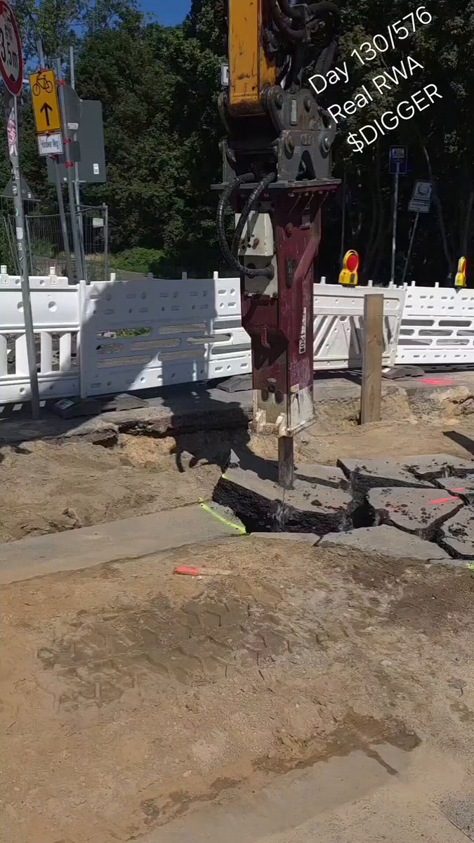 The image shows a construction site with a large excavator digging into the ground. The excavator is positioned near a white fence, which has orange cones placed around its base for safety purposes. There are also two workers present at the site, one of whom is operating the excavator. In addition to these elements, there's a sign that reads "Day 130 of 576. New week, new hole, new plan to execute, no shortcuts."