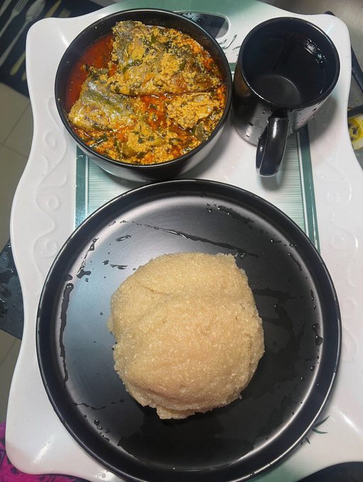 A top-down view of a meal on a white tray shows a bowl of fish stew in red sauce, a black mug, and a round serving of fufu or dough on a black plate.