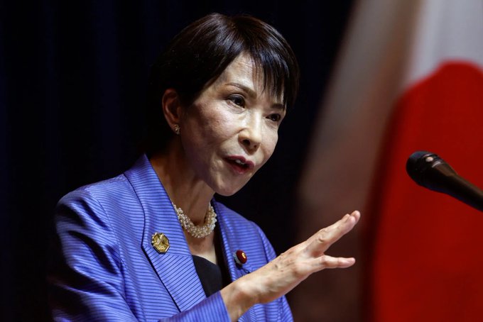 The image shows an older woman wearing a blue blazer and pearl necklace, standing at a podium with her hand outstretched as if she's giving a speech or presentation. The background features a flag from Japan, which suggests that this event might be taking place in Japan. The caption provided after the image reads "NOW: BANK OF JAPAN HIKES RATES BY 25BPS TO THE HIGHEST LEVEL SINCE 19950". This indicates that the woman is speaking at an event related to banking or finance, possibly discussing the Bank of Japan's monetary policies and their impact on the Japanese economy.