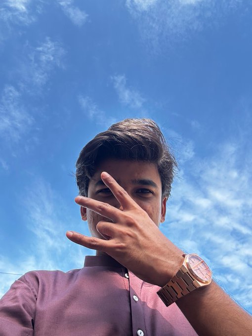 A low-angle selfie of a young man with dark hair, wearing a maroon shirt and a gold watch, holding his hand up against his face with fingers spread, set against a bright blue sky with wispy clouds.