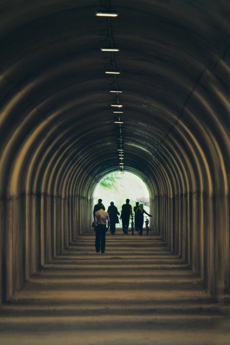 A view through a long, arched tunnel with overhead lights receding into the distance, where silhouetted figures walk toward a bright exit.