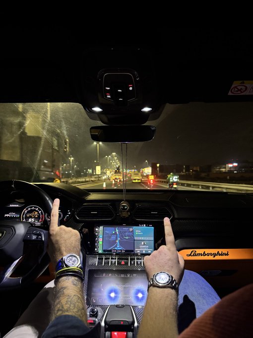 A first-person view from inside a Lamborghini at night, with the driver's tattooed arms raised and middle fingers up toward the windshield showing highway traffic.