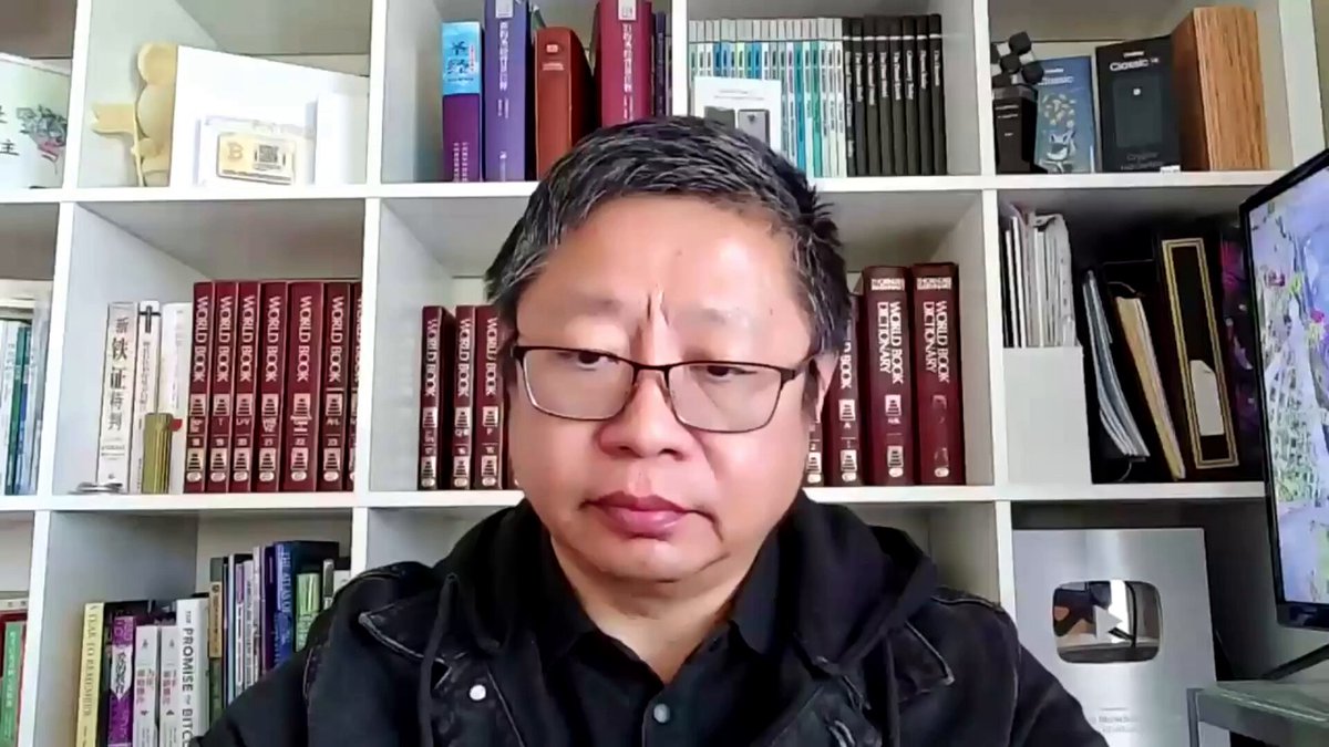 The image shows a man wearing glasses sitting at a desk with bookshelves behind him, filled with various books. The man appears to be looking directly into the camera while seated. There are no other people or objects visible in the image, but the presence of numerous books suggests that he might be an avid reader or involved in some form of academic or research work.