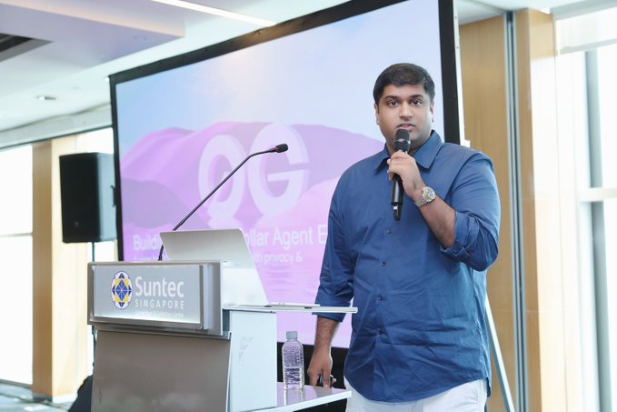A man in a blue shirt speaks into a microphone while standing at a podium during a 'Suntec Singapore' event, with a presentation screen displaying 'OG' behind him.