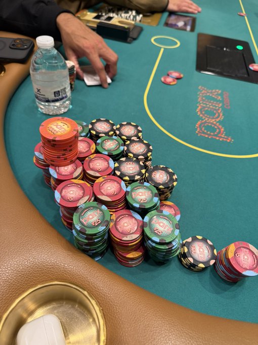 A large stack of colorful poker chips sits on a green felt casino table with yellow markings, alongside a water bottle, a smartphone, and a player's hand reaching for cards.