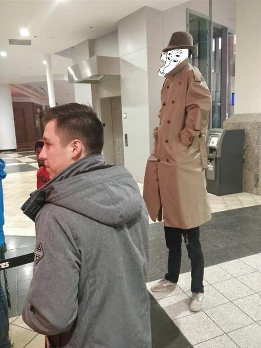 A man in a long tan trench coat, brown hat, and white shoes stands tall in a lobby, while a shorter man in a grey jacket stands in the foreground; the tall man's face is obscured by a cartoon meme sticker.