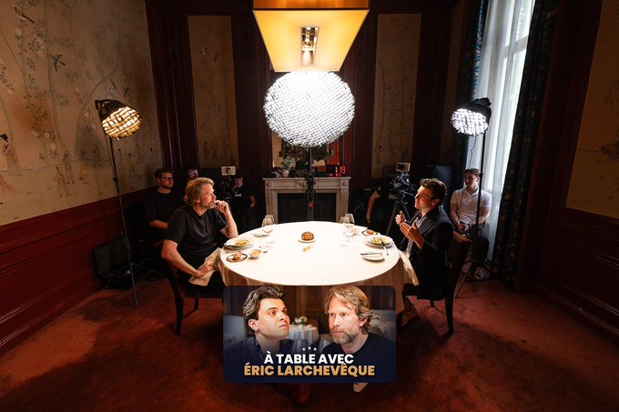A behind-the-scenes photo of a TV show set titled 'À TABLE AVEC ÉRIC LARCHEVÊQUE' showing two men seated at a dining table under studio lights, with crew members and filming equipment visible.