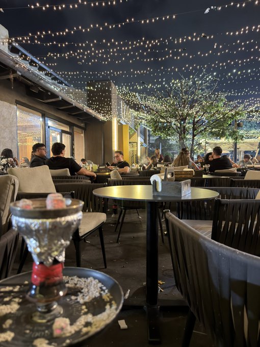 An evening photograph of an outdoor restaurant or bar patio decorated with extensive string lights overhead, featuring wicker chairs, round tables with patrons dining and socializing, with a small decorative goblet visible in the foreground.