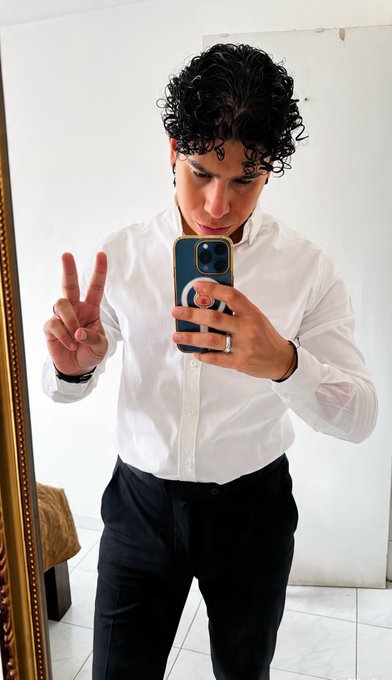 Young man in a white dress shirt and black pants taking a mirror selfie with a blue iPhone while making a peace sign.