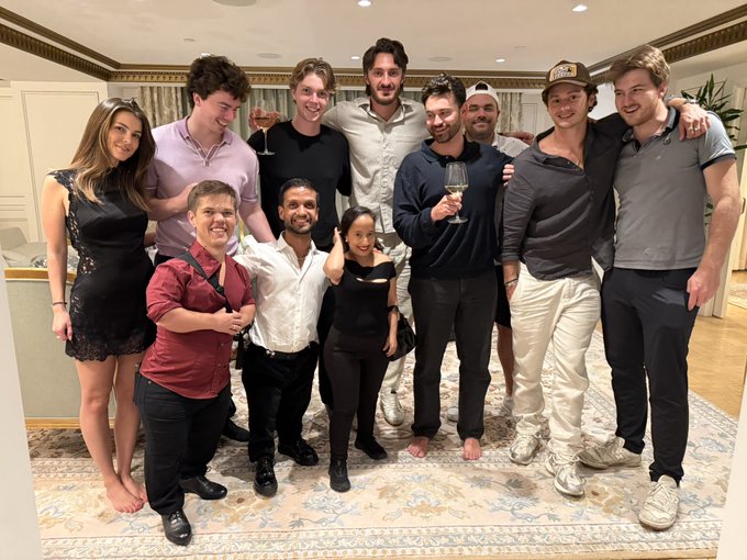 A group of ten young adults pose closely together in a carpeted hotel suite, smiling and raising a wine glass in a celebratory toast. The casual gathering fits the post's tone, marking a relaxed "summer baby" welcome into the $SYN Waldwarf Hysteria community.