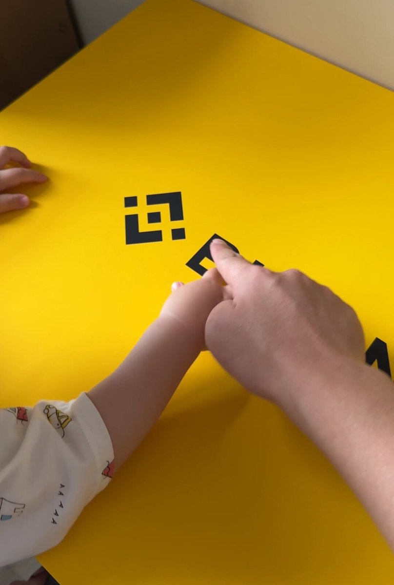 The image shows a person's hand pointing at a black square with a letter "B" on it, which appears to be part of an educational game or activity involving letters. The background features a yellow surface, possibly a table or desk, where the game is taking place. There are also other people in the scene, but they are not the main focus of the image.