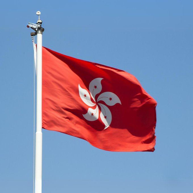The Hong Kong flag, featuring a white bauhinia flower with red stars on a red field, waves against a clear blue sky. The image visually represents Hong Kong as the article discusses the region's plans to pilot tokenized Exchange Fund Bills by the end of 2026.