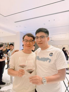 Two young men wearing glasses pose together at an indoor event, each holding a glass of red wine, with one wearing a white T-shirt with blue text.