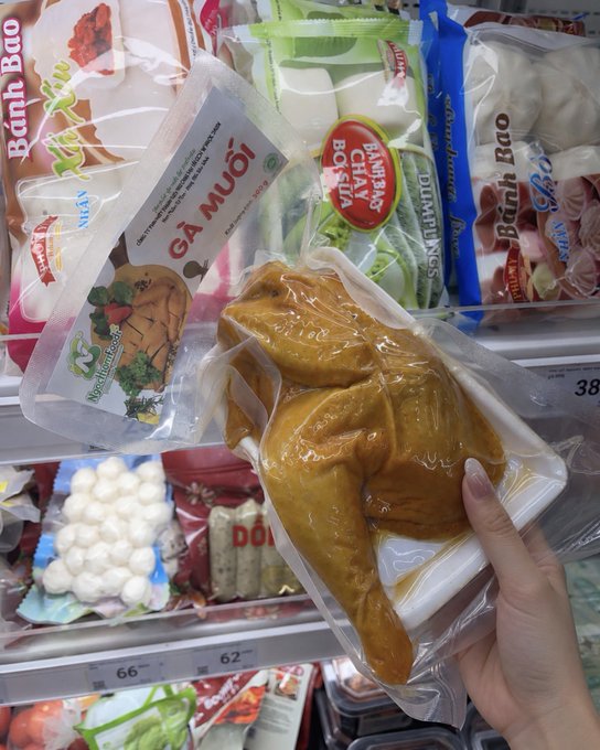 A hand holds a vacuum-sealed package of a whole boiled chicken (labeled 'GÀ MUỐI') in a refrigerated store section alongside Vietnamese food items including bánh bao and dumpling wrappers.