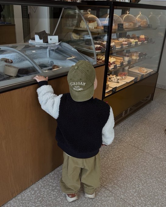A young child wearing a black fleece vest, khaki pants, and a "Michigan State Original" baseball cap stands looking into a bakery display case filled with cakes and pastries.