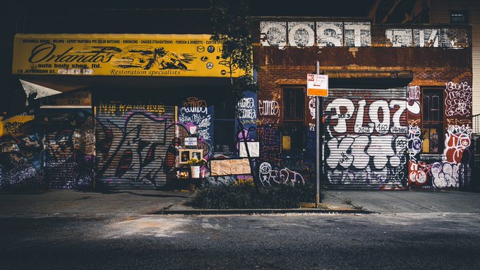 A nighttime street scene shows a building with a yellow sign reading 'Orlando's' for restoration services, while the storefront below is covered in colorful graffiti including tags like 'CLUTCH' and 'WASTE'.