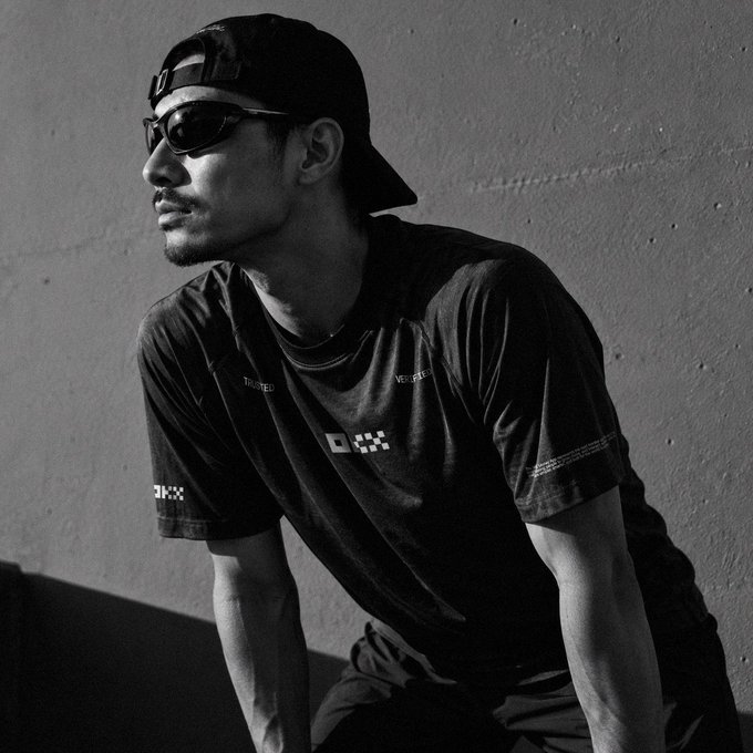 A black-and-white portrait of a young man wearing sunglasses, a backwards cap, and a dark graphic t-shirt.