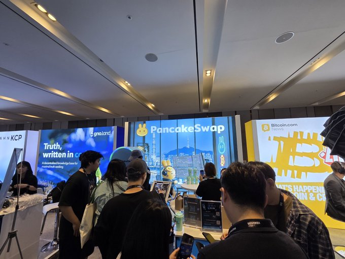 A crowded tech conference booth area featuring large screens displaying sponsors 'PancakeSwap' and 'Bitcoin.com News', with attendees mingling and using their phones.