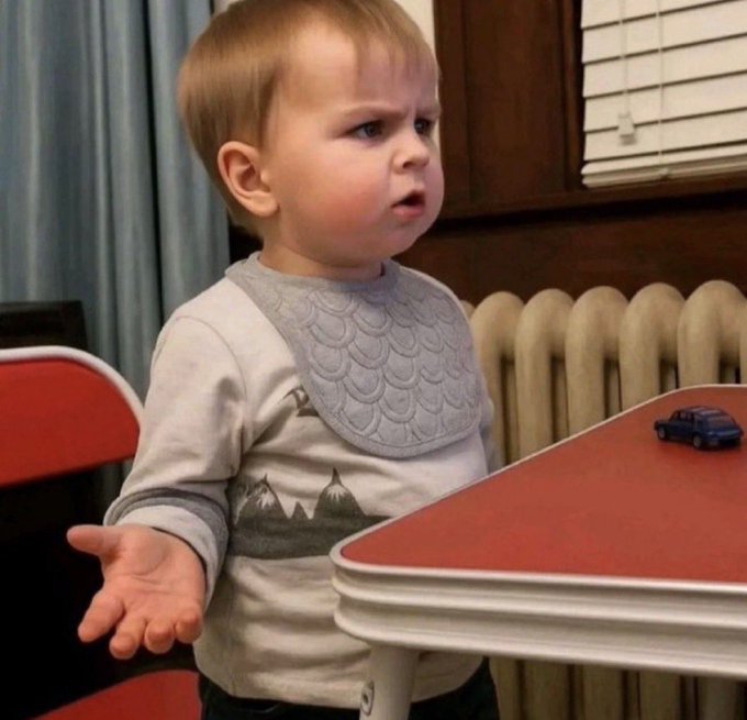 A young toddler wearing a grey and white patterned bib stands at a red retro-style table, looking confused or displeased next to a small blue toy car.