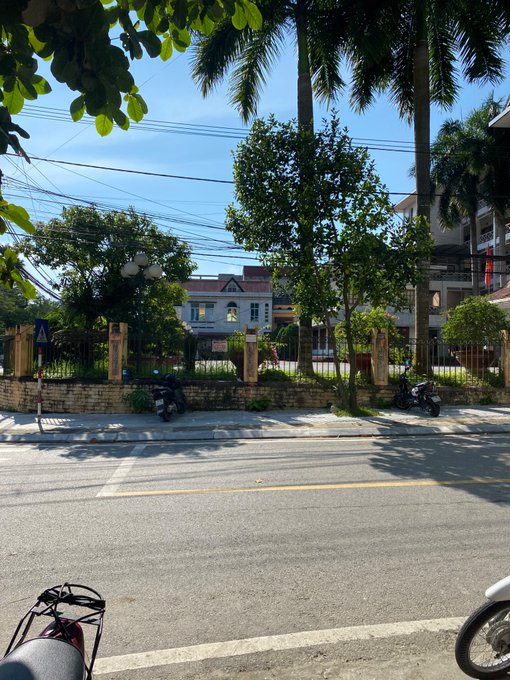 A sunny street view shows a road with motorbikes parked along a stone wall, with palm trees and a white building labeled 'Khu Biệt thự' visible across the street.