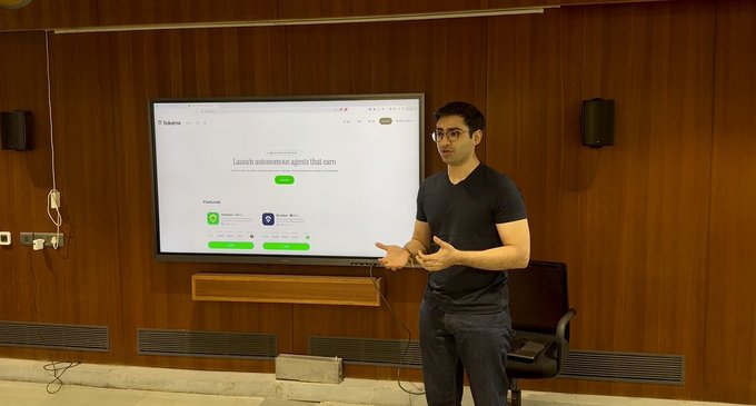 A man in a black shirt and glasses presenting in front of a large screen displaying the Sukonto app's autonomous agent launch page.
