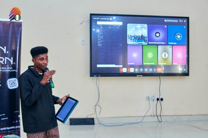 A young man in a black hoodie holding a microphone and a tablet, presenting in front of a wall-mounted TV displaying a digital interface, with a promotional banner visible on the left.