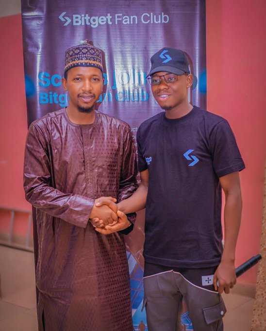Two men pose together shaking hands in front of a banner for the 'Bitget Fan Club'; one wears traditional maroon attire and a cap, while the other wears a black Bitget t-shirt, cap, and glasses.