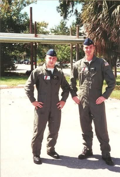 Two men in green military flight suits stand outside a covered structure, with the younger man on the right wearing glasses and a rank chevron.