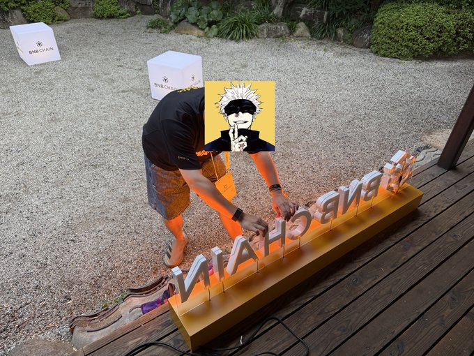A person is repairing fallen illuminated letters that spell "MIAHOEVANGE" on a wooden display at a BNBCHAIN venue, gluing the letters back into place on the gold base. The scene illustrates the caption's description of someone quietly doing good deeds by fixing the fallen light signage at the event.