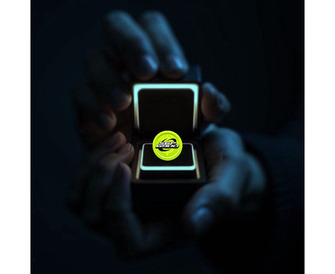 Two hands in dim lighting hold open a small jewelry box containing a glowing yellow circular token with the 'GRASS' logo on it.