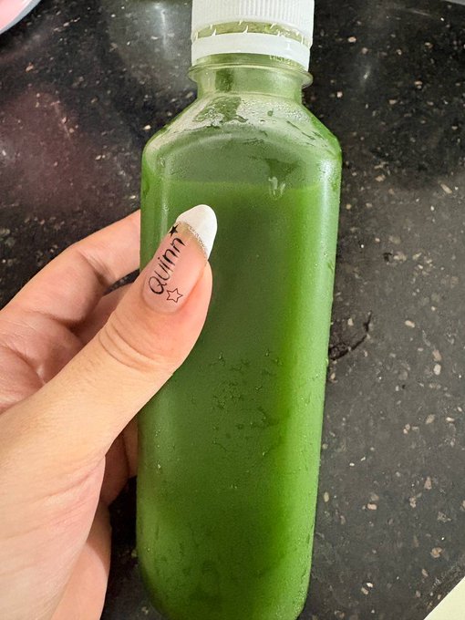 The image shows a person's hand holding a green smoothie bottle with a white cap. The text "qui" can be seen on the bottle as well, possibly indicating that this is a detox smoothie or a similar product. The background of the photo appears to be a kitchen countertop.