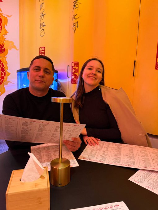 The image shows a man and woman sitting at a table, looking at menus or reading papers while smiling for the camera. The background features a yellow wall adorned with Chinese characters that read "福", which translates to "Chinese". There is also a gold-colored lamp on the table next to them.