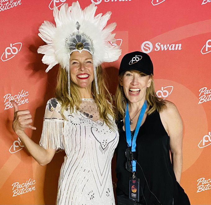 Two women posing in a photo booth at a Pacific Bitcoin event with a red backdrop; one wears an elaborate white feathered headdress and beaded dress, while the other wears a black cap and shirt with a 'STAFF' lanyard.