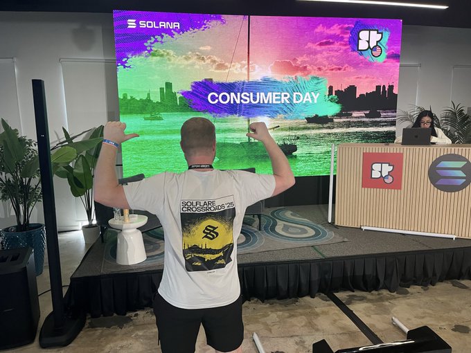 A person in a white 'SOLFLARE CROSSROADS '25' t-shirt flexes their arms while facing a large screen displaying 'SOLANA CONSUMER DAY' and the 'SP' logo at a conference.