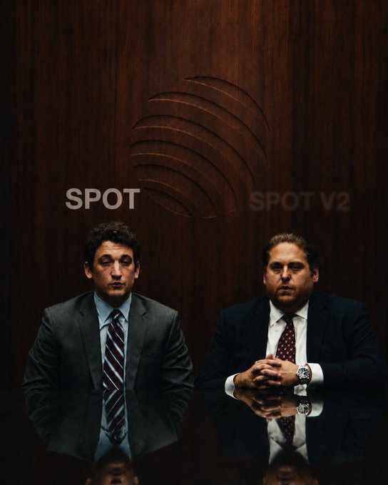 Two suited men sit at a reflective table labeled "SPOT" and "SPOT V2" against a dark wood backdrop, evoking a corporate interview setting reminiscent of the Bloomberg "Big Decisions" segment. The "V2" implies a new, accessible version of something previously reserved for experts, aligning with the post's theme of democratization and a major upcoming announcement.