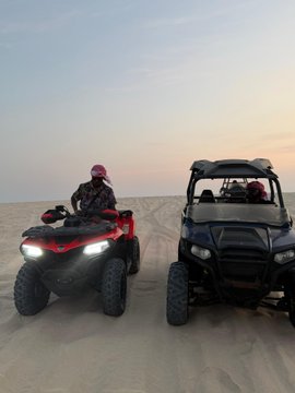 Two people sit on ATVs—a red Honda on the left and a black side-by-side UTV on the right—on sand dunes at sunset.