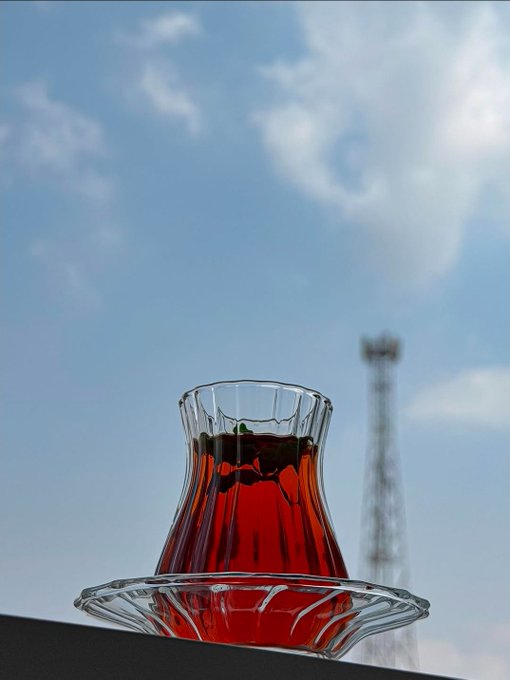 A tulip-shaped glass of red tea sits on a saucer against a bright blue sky with scattered clouds, with a blurred transmission tower in the background. The setting suggests a relaxed afternoon, aligning with the post's "GOOD AFTERNOON" greeting while the imagery of everyday accessibility contrasts with the ambitious vision of decentralized quantum computing infrastructure described in the message.