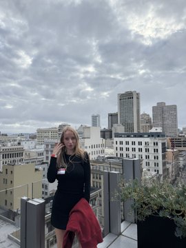 A blonde woman in a black dress stands on a rooftop terrace talking on her phone, with a city skyline and cloudy sky in the background.