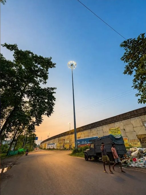 A street scene at dusk featuring a tall light pole, a stone wall with posters and signs, a parked truck, and two people standing near piles of goods along the roadside under a clear blue sky.