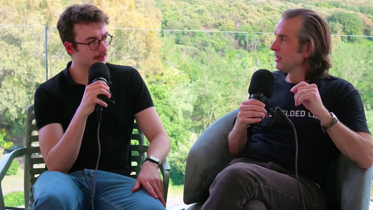 The image shows two men sitting next to each other with microphones in their hands, suggesting they are participating in a podcast or interview. The background features a view of trees, indicating that they might be recording from an outdoor location such as a park or garden. The focus on these elements helps convey the idea that the conversation is centered around a specific topic related to the environment and sustainability.