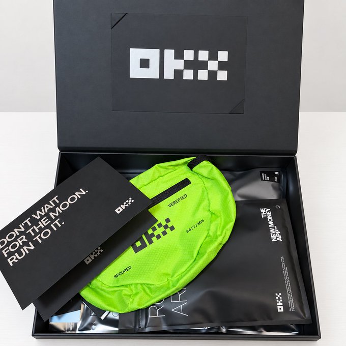 An open black OKX-branded box contains neon green merchandise, including a fanny pack labeled 'VERIFIED' and 'SECURED' with number 1477389, a card reading 'DON'T WAIT FOR THE MOON. RUN TO IT.', and a package labeled 'THE NEW MONEY APP'.