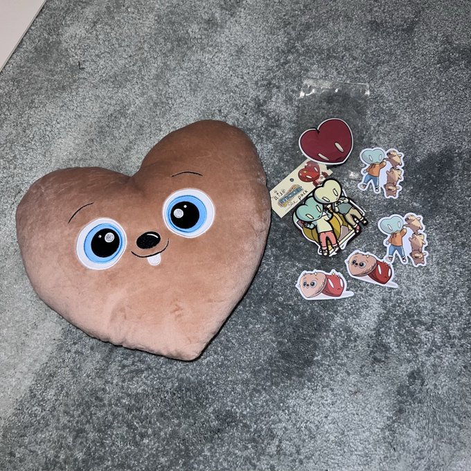A plush heart-shaped pillow with cartoon eyes and a mouth sits next to a set of small stickers featuring cute characters.