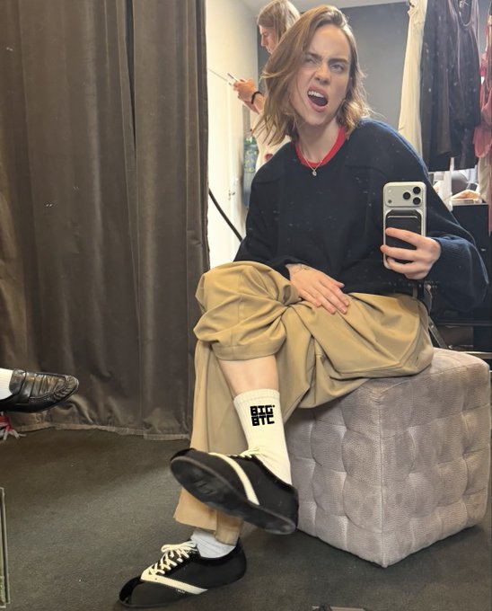 A person takes a mirror selfie while sitting on a gray ottoman, wearing a navy sweater, khaki cargo pants, and white socks featuring 'BIG BTC' text.