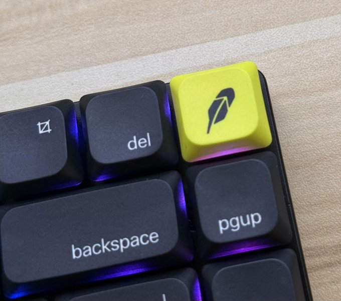 A close-up of a backlit mechanical keyboard shows a yellow keycap featuring a quill/feather logo, positioned above standard keys like 'del,' 'backspace,' and 'pgup.'