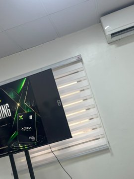 A photo taken from a low angle showing a wall-mounted air conditioning unit above a flat-screen TV displaying a 'XORA' branded graphic, with blinds on the window behind it.