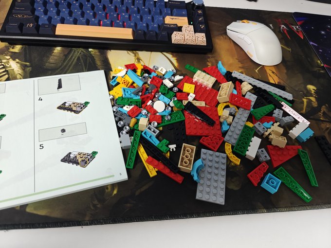 A pile of colorful Lego pieces scattered on a desk next to an open instruction manual, with a mechanical keyboard and white computer mouse visible in the background.