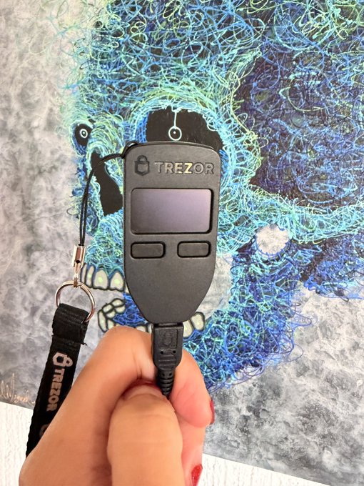 A hand holds a Trezor hardware wallet with its branded lanyard against an artistic blue abstract painting, illustrating the user's mention of an old Trezor device that they're planning to upgrade.