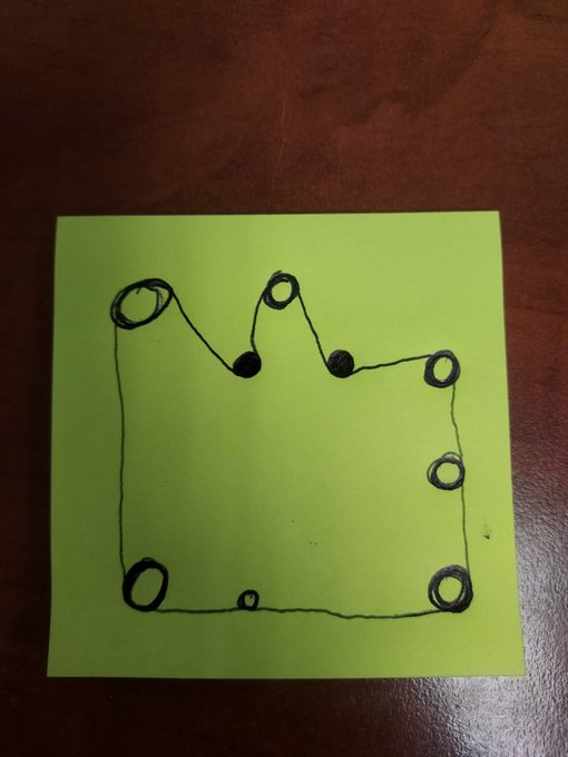 A hand-drawn sketch in black marker on a bright green sticky note depicting a crown-like shape with circular nodes connected by lines, sitting on a dark wooden surface.