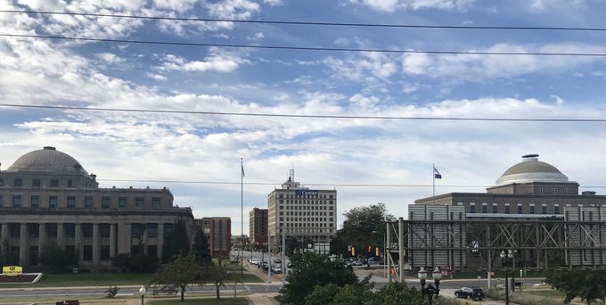 A daytime cityscape featuring two domed government-style buildings on either side of a street, with a blue and white flag (likely Chicago's) flying, under a sky filled with scattered clouds.
